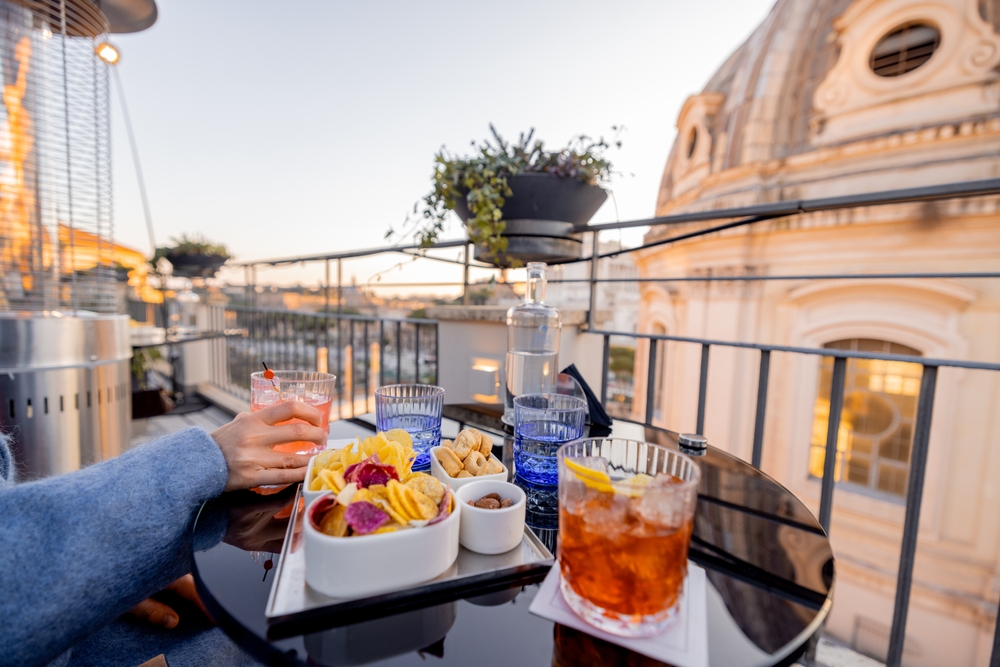 A kép egy elegáns aperitivot ábrázol koktélokkal és rágcsálnivalókkal egy római tetőteraszon, naplementekor.  - Z(s)eppelin utazás